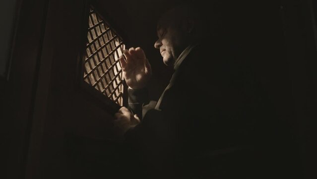 Christian church monk crossing and praying in confessional booth low angle view. Religious man sitting near confession window. Asking for forgiveness concept