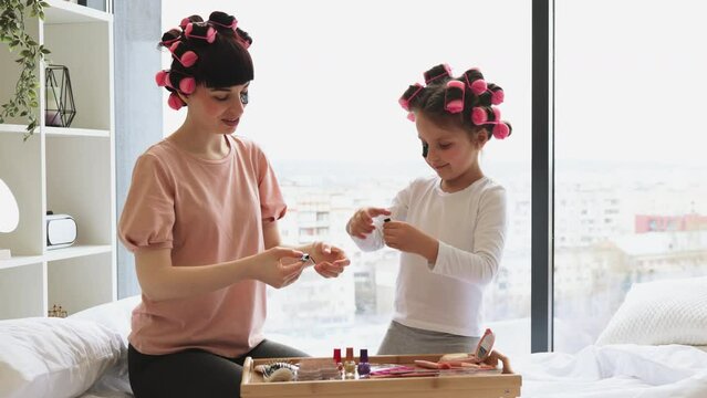 Caucasian family mother and daughter perform cosmetic procedures at the weekend while sitting on the bed. Young woman and cute girl take eye patches from cosmetic table.