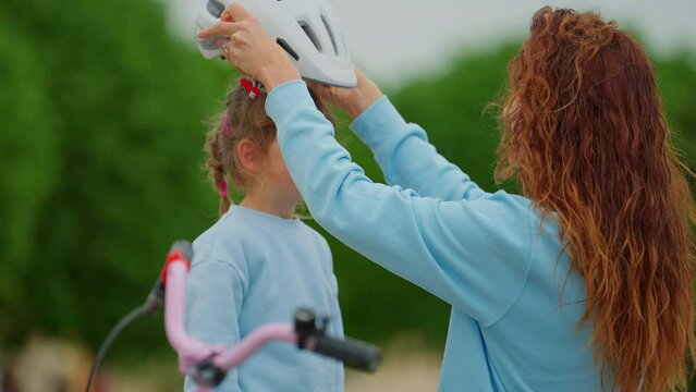 Loving mother put on sport helmet to little cute kid daughter before spin bicycle on street. Little girl wear protective halmet for health care and safety