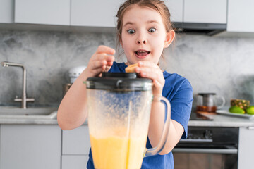 Portrait of a crazy little girl with a blender in the kitchen. Cheerful little girl makes a fruit...