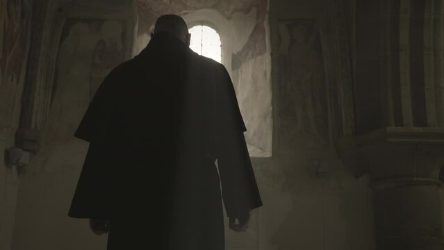 Bald Christian monk in robe silhouette standing in front of sunlit window in church back view medium shot. Religious prayer in monastery. Christianity concept