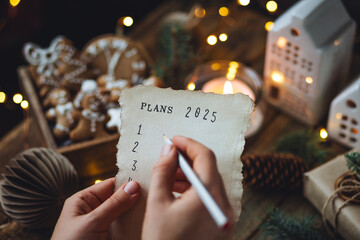 Writing new year resolutions 2025 by hand in December on New Year's Eve. Plan, goals, ideas, projects. Cozy home holiday atmosphere, Christmas lights, festive decor, homemade gingerbread cookies