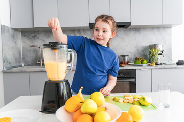 Portrait of a cute little girl making a fruit cocktail at home in the kitchen. A girl puts fruit into a blender. Selected focus