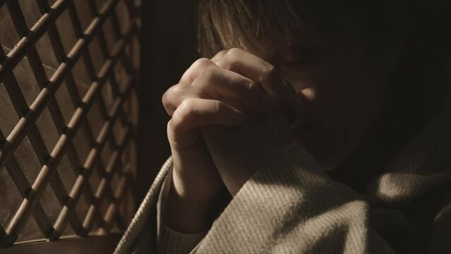 Religious woman praying with clasped hands near confessional booth window. Female parishioner speaking, asking for forgiveness during confession in church close up