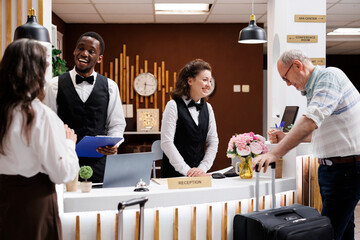 Retired senior couple arrive at hotel, assisted by smiling multiethnic staff at front desk....