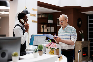 Caucasian retired old man signs reservation documents at hotel front desk as african american...