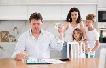 Wistful father sharing his money on the table with wife, their little son and daughter standing by mother in the kitchen