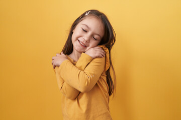 Adorable hispanic girl with a joyful smile standing and hugging herself on isolated yellow background, exuding confidence and positive self love - obrazy, fototapety, plakaty