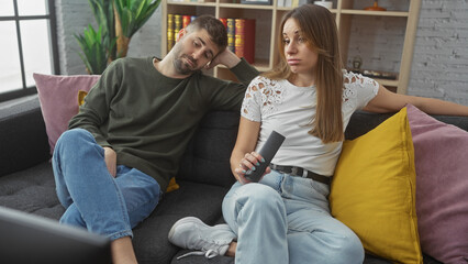 A disinterested woman and a man lounging on a sofa in a modern living room, showing boredom with a remote control in hand.