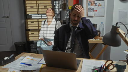 Stressed man and contemplative woman detectives working late in cluttered office, surrounded by...