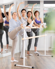 Enthusiastic senior woman participating in stretching exercises at barre with group of active women...