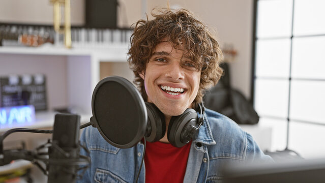 A smiling young man with curly hair wearing headphones in a recording studio.