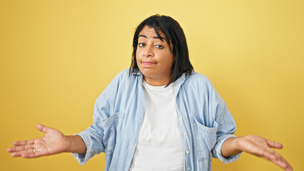 A perplexed hispanic woman shrugs against a yellow wall, embodying uncertainty and confusion in a...