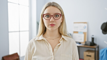 Portrait of a serious young woman with blonde hair and red glasses standing in a modern office...