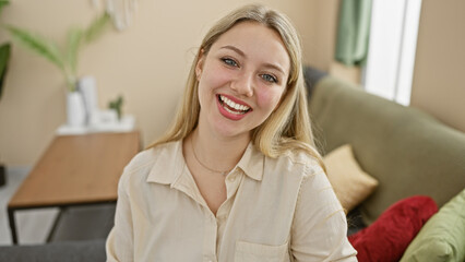 Smiling blonde woman in a casual beige shirt posing comfortably at her modern furnished living room.