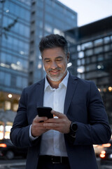 Smiling mature hispanic senior business man using app, holding smartphone cellphone at office...