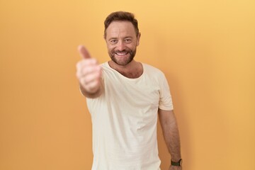 Middle age man with beard standing over yellow background smiling friendly offering handshake as...