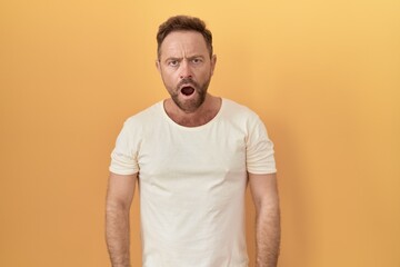Middle age man with beard standing over yellow background in shock face, looking skeptical and...