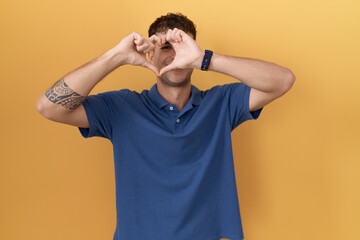 Young hispanic man standing over yellow background doing heart shape with hand and fingers smiling looking through sign