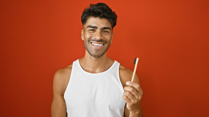 A cheerful young hispanic man with a beard poses in a sleeveless shirt against an isolated red...