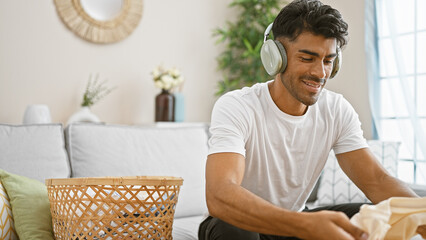 Hispanic man folding laundry at home while listening to music on his headphones captures a relaxed...
