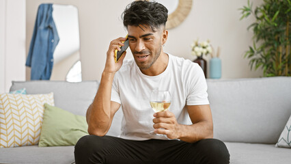 Hispanic man in white shirt talking on phone holding wine glass indoors