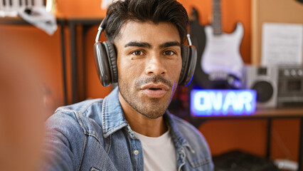 Handsome hispanic man with beard wearing headphones in a music studio with guitar and 'on air' sign