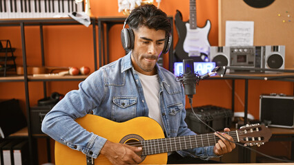 Handsome hispanic man playing guitar and singing in a music studio with recording equipment