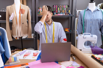 Young hispanic man dressmaker designer using laptop surprised with hand on head for mistake,...