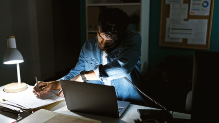 A young hispanic man works late in an office, writing under a lamp with a laptop and phone.