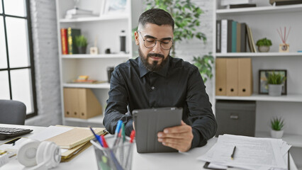 Bearded man using tablet in modern office with headphones, papers, and shelving.
