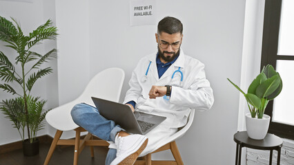 Bearded man in lab coat checks watch while working on laptop in modern clinic lobby with free wifi...