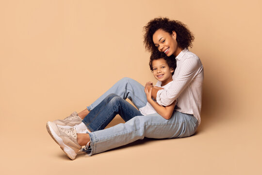 African American woman is sitting on the ground with a child beside her. The woman appears to be engaging with the child in conversation or play, both seem relaxed in their interaction.