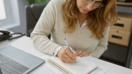 A professional woman concentrates on writing notes at her office desk surrounded by laptop and...