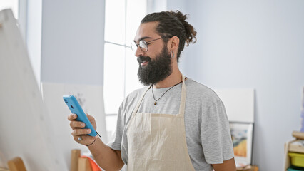 Hispanic man with beard wearing glasses using smartphone in art studio indoor setting