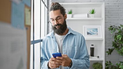 Handsome hispanic man with beard using smartphone in modern office, plants in background, embodying...