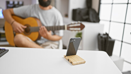Hispanic man playing guitar indoors with smartphone recording on white table.