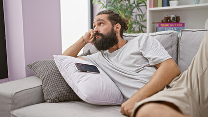 Thoughtful bearded man lounging on couch with smartphone, expressing relaxation and comfort in a...