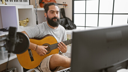 Hispanic man with beard playing guitar indoors, facing a computer on studio desk with microphone.