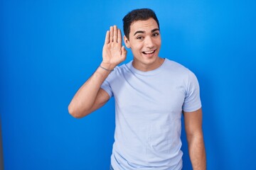 Young hispanic man standing over blue background waiving saying hello happy and smiling, friendly...