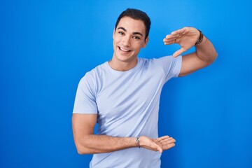 Young hispanic man standing over blue background gesturing with hands showing big and large size sign, measure symbol. smiling looking at the camera. measuring concept.