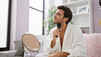 Hispanic man in white bathrobe grooming with electric trimmer in modern living room.