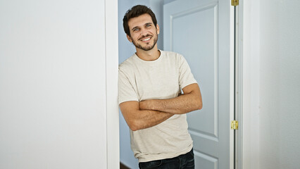 Handsome young hispanic man with a beard standing arms crossed in a casual home interior