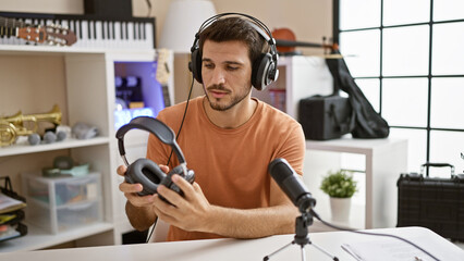 Handsome hispanic man examining headphones in a modern music studio setting, surrounded by...