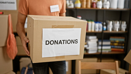 Hispanic man carrying donation box in a cluttered volunteer warehouse filled with shelves and supplies.