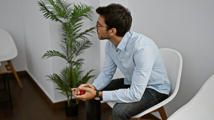 A young hispanic man with a beard squeezes a stress ball indoors, sitting in a room with a white...