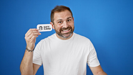 Smiling bearded man in a white t-shirt shows a gift card against a vibrant blue background,...