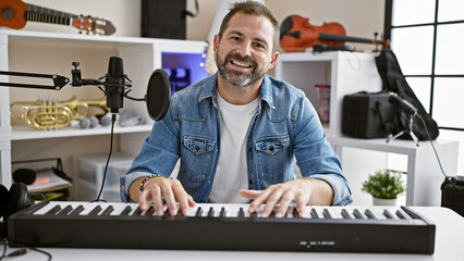 A smiling hispanic man with grey hair playing a keyboard in a music studio, surrounded by guitars...