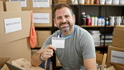 Smiling middle-aged man displaying id in a donation center with boxes labeled donations in...