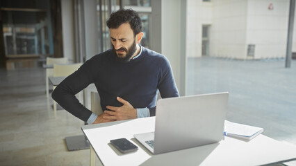 A middle-aged bearded man in discomfort at his modern office workplace indoors, with a laptop and smartphone on the desk.